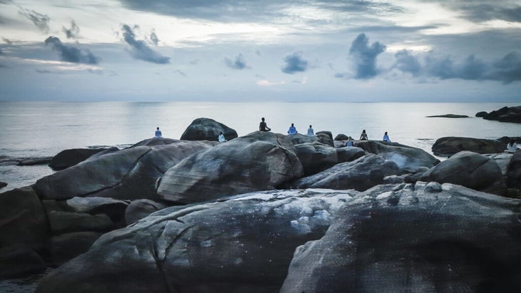 people sitting on a rock formation in easy pose overlooking the ocean with a cloudy sky at a wellness retreat